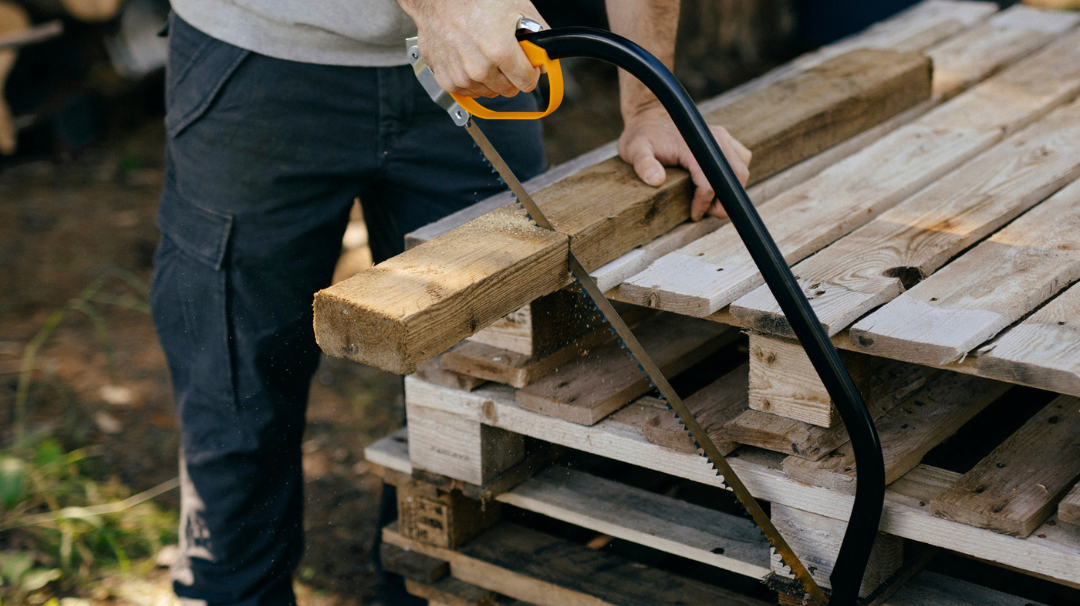 Bow saw being used to cut through a thick wooden beam resting on stacked timber pallets.