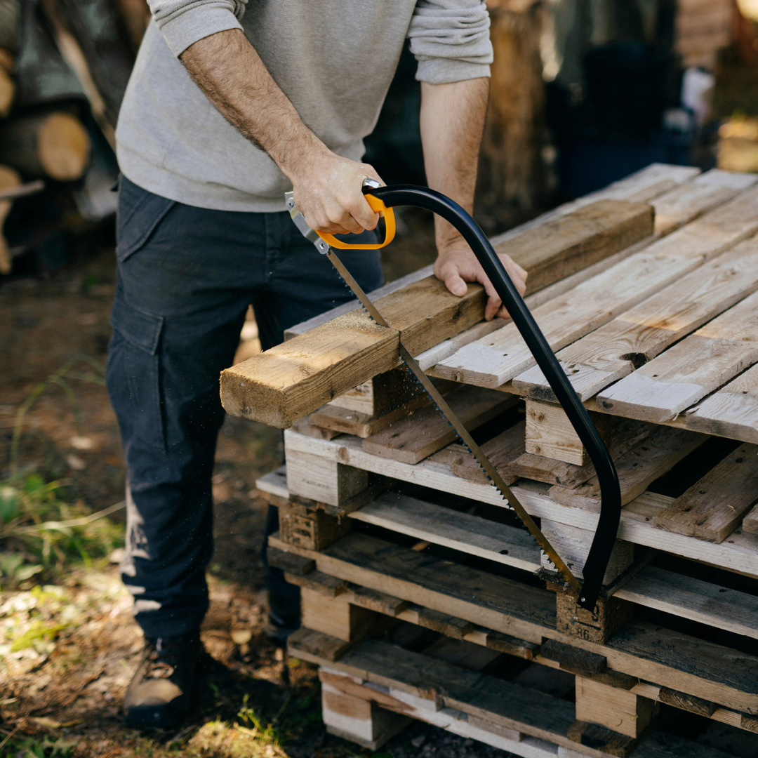 Bow saw being used to cut through a thick wooden beam resting on stacked timber pallets.