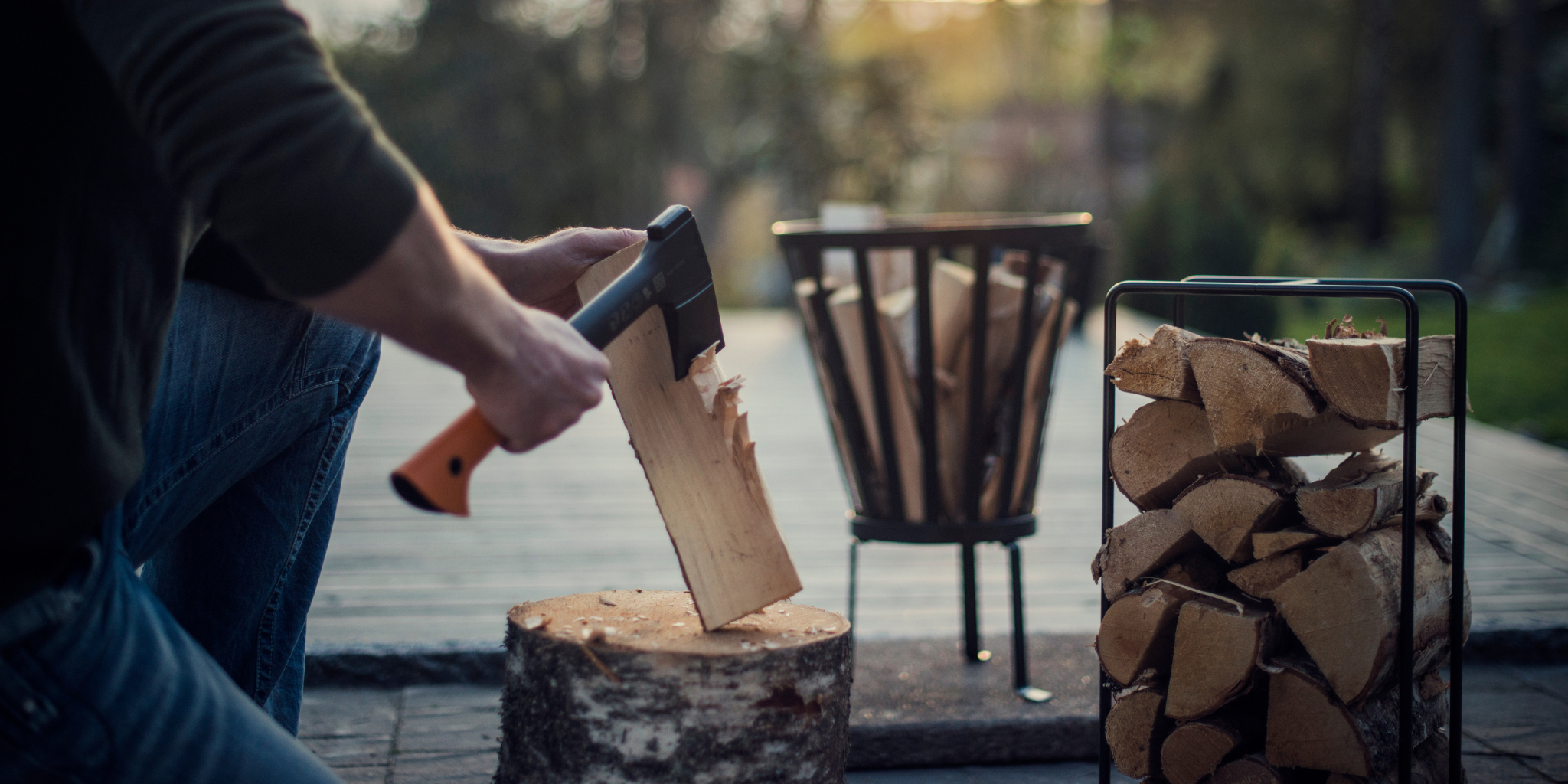 Person using a splitting axe to split firewood beside a stacked wood rack and outdoor fire pit.